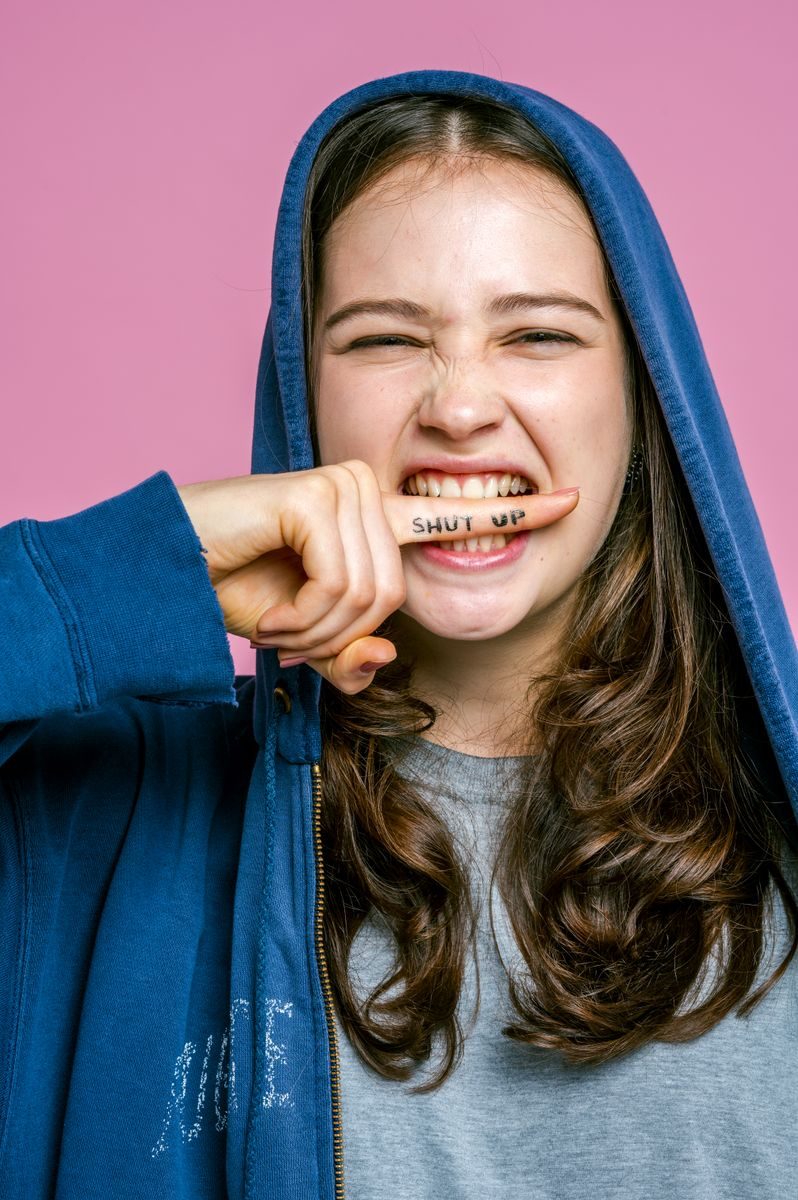 Teen girl playfully displays 'shut up' message on finger against pink background.