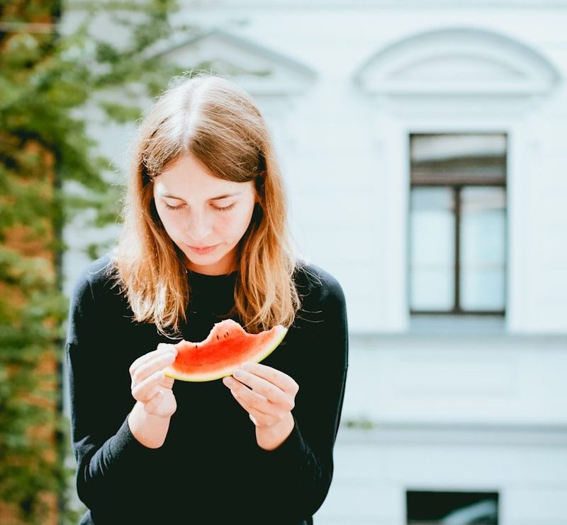woman holding slice of watermelon while sitting on concrete bench outdoor