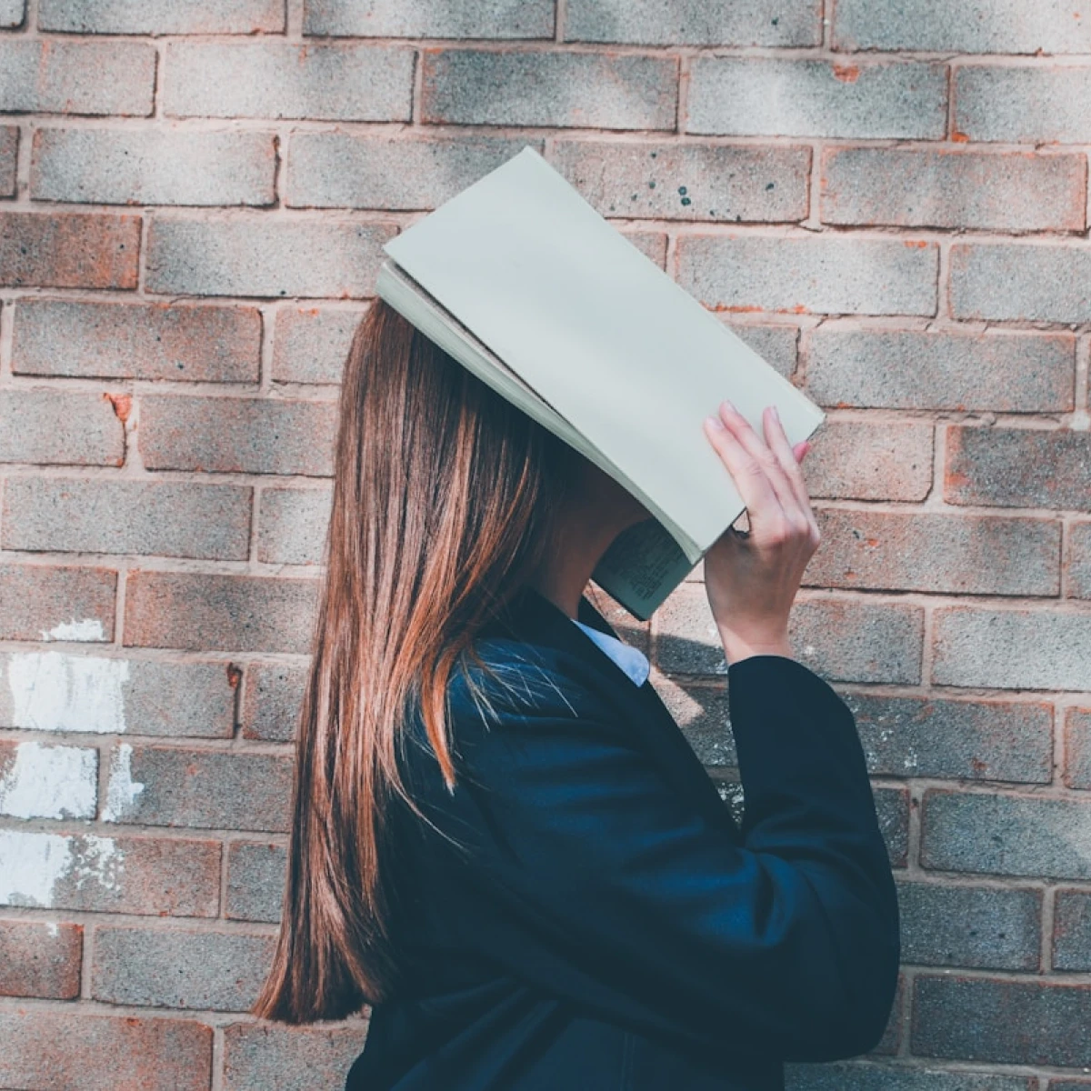 Pupil-covering-face-with-books-in-front-of-brick-wall