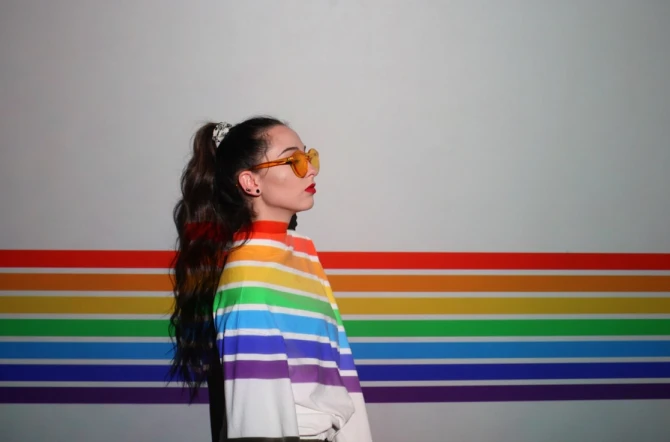 girl with sunglasses and rainbow sweater standing side on against a rainbow wall