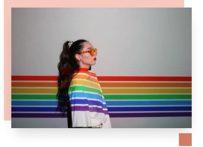 girl with sunglasses and rainbow sweater standing side on against a rainbow wall