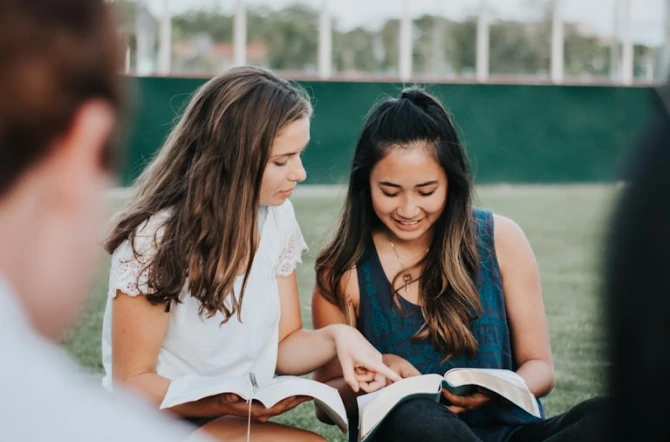 Two girls sitting next to each other reading books with two other friends in the foreground blurred out of the shit