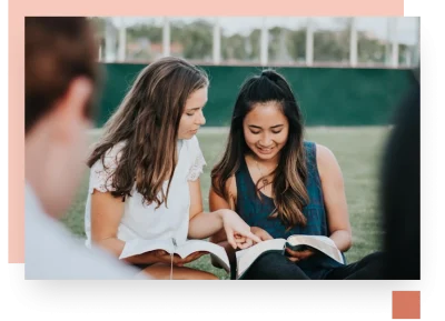 Two girls sitting next to each other reading books with two other friends in the foreground blurred out of the shot