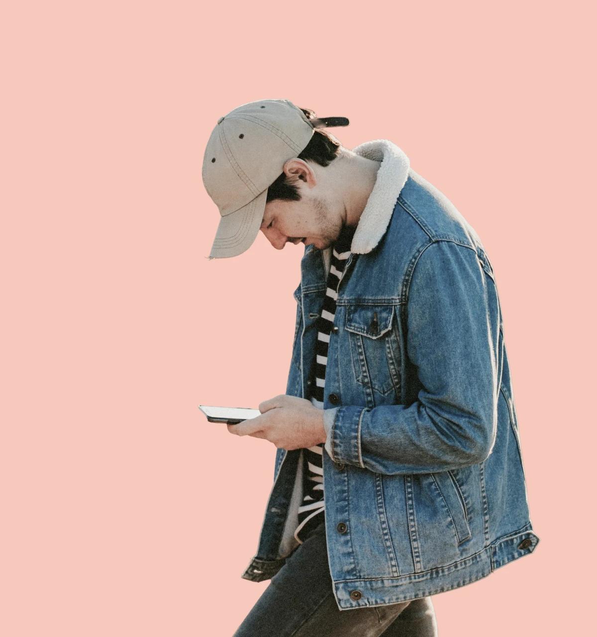 Teen wearing cream baseball cap and blue denim jacket looking down at phone on plain apricot background